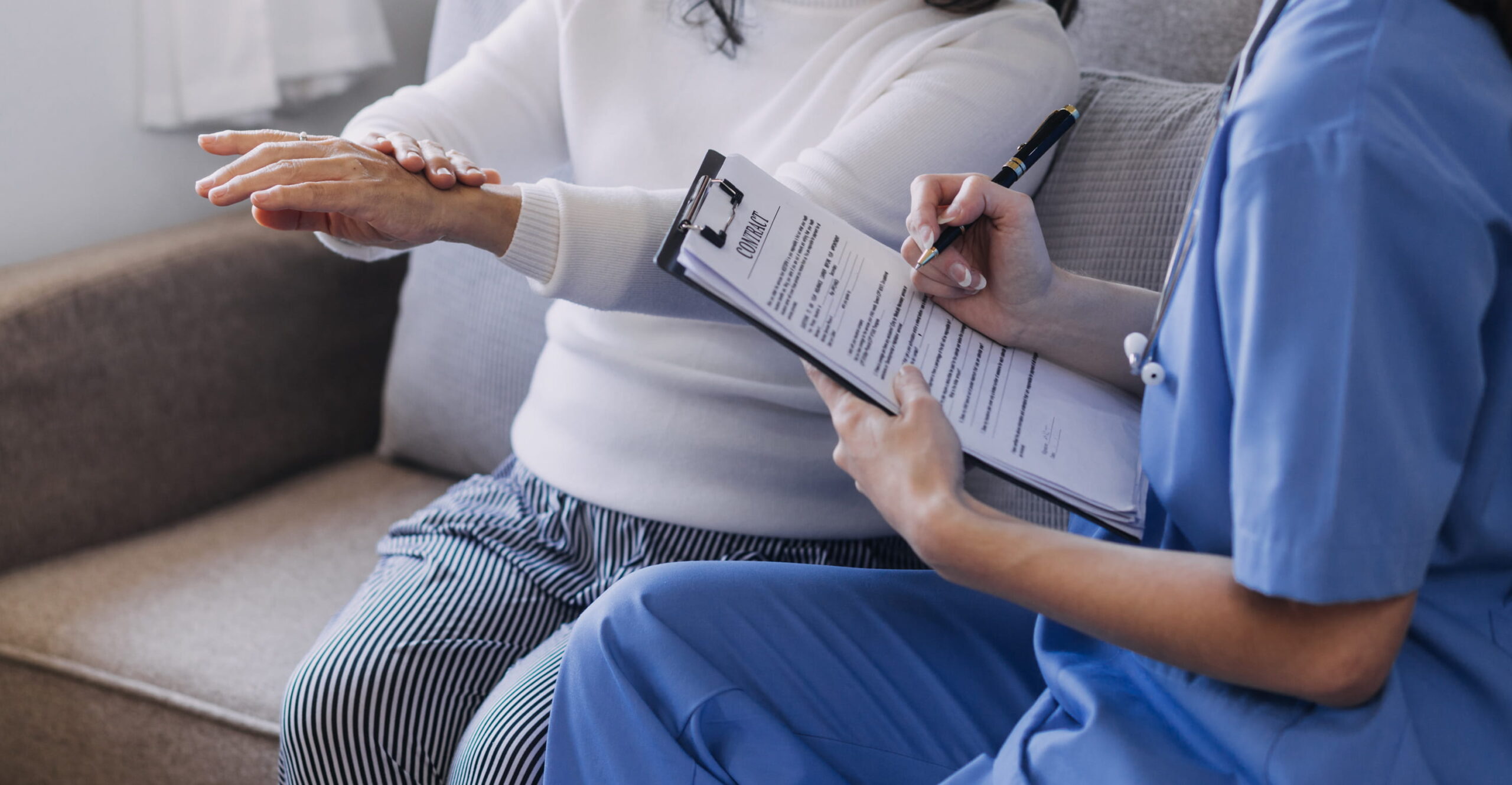 Pflegegrad-Antrag Schritt für Schritt: Eine Pflegefachkraft bespricht bei einem Hausbesuch Unterlagen mit einer älteren Frau und macht sich Notizen zur Pflegesituation.